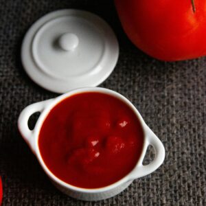 Close-up of tomato sauce in a ceramic bowl with fresh tomatoes on textured fabric.