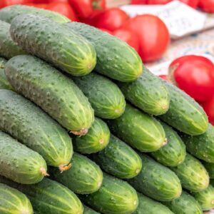 A stack of fresh cucumbers displayed at an outdoor farmers market with ripe tomatoes.