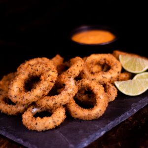 Vibrant orange salad dressing and fried brown round onion rings composing with slices of lime on wooden table in daylight