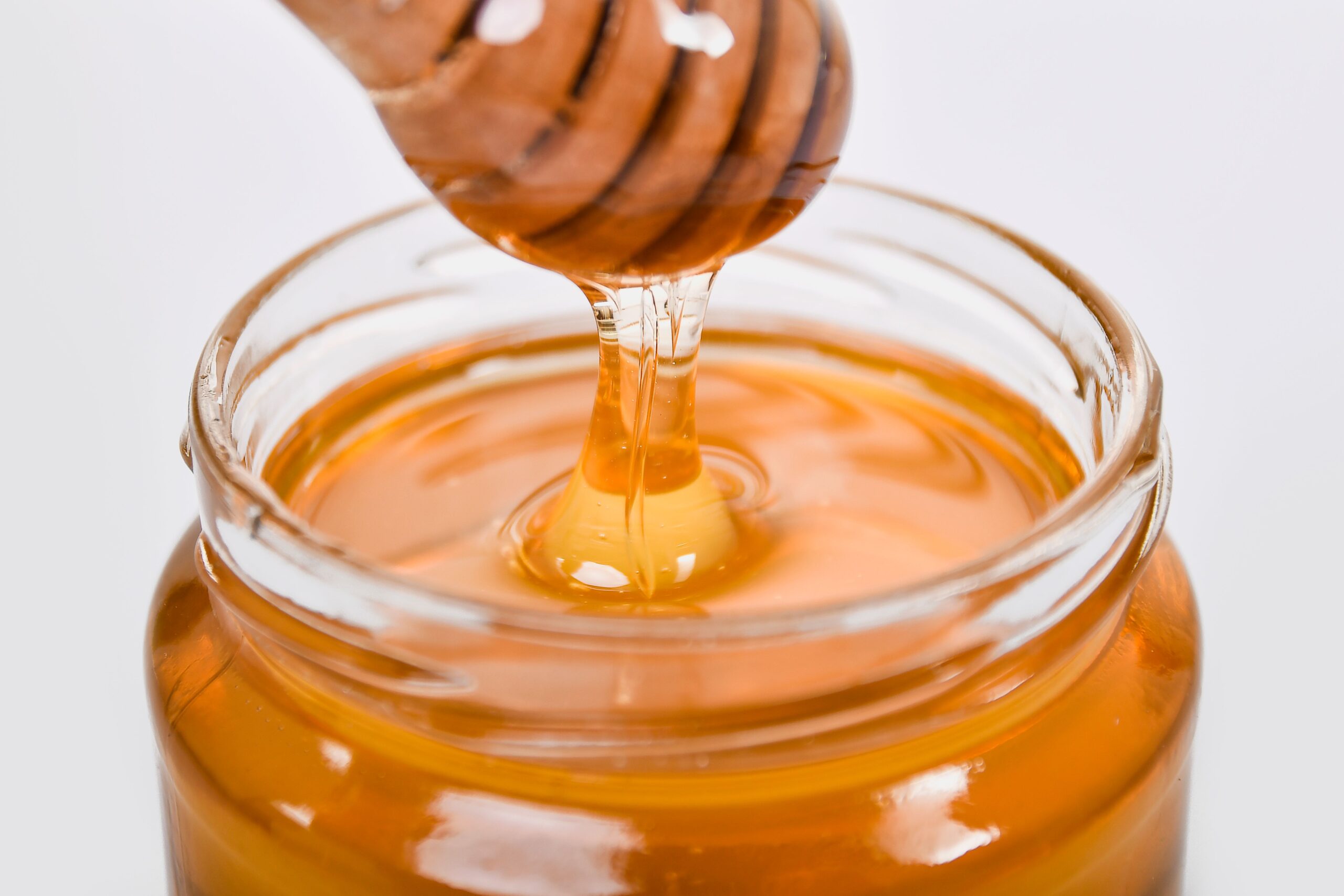 pexels photo 5634206 5634206 Close-up of honey flowing from a wooden dipper into a glass jar on a white background.