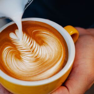 Unrecognizable professional barista pouring milk from pitcher into yellow cup with latte with creative pattern while working in coffee house
