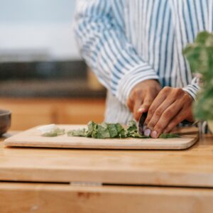 Crop anonymous ethnic male in striped shirt cutting fresh spinach on chopping board while cooking at home